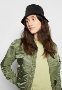 Casaco bomber de cetim verde com fecho de correr, detalhes em dourado e punhos canelados, usado sobre uma camisa verde clara. Um chapéu de balde preto completa o visual.
