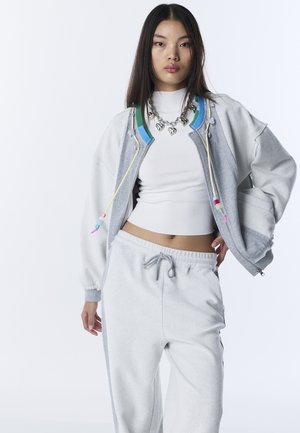 Jonge vrouw die een wit crop-topje draagt, een grijs-wit zip-up hoodie met kleurrijke kralen, grijze joggingbroek en een dikke zilveren ketting met hartvormige bedels.