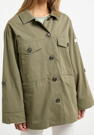 Femme portant une veste oversize vert olive à boutons avec poches à rabat sur la poitrine et manches longues par-dessus un haut vert clair.