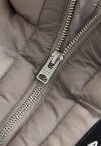 Grå puffjacka med ett inre som har en silverdragkedja med en märkesdragflik och quiltad textur. Slät tyg med lätt lyster.