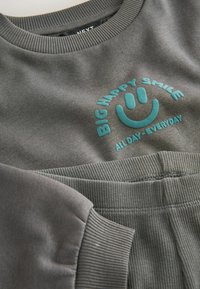 Sweat gris et pantalon avec poignets côtelés, avec texte turquoise et visage souriant : « GRAND SOURIRE HEUREUX TOUTE LA JOURNÉE - TOUS LES JOURS » sur la poitrine.
