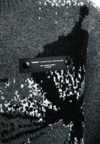 Suéter de punto en tonos grises y negros que presenta un patrón texturizado y una etiqueta de cuero negro con texto en relieve y botones a presión.