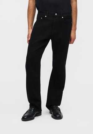 Mężczyzna ubrany w czarne jeansy o prostych nogawkach i czarne skórzane mokasyny, stojący z jedną ręką swobodnie opuszczoną wzdłuż ciała na jednolitym białym tle.