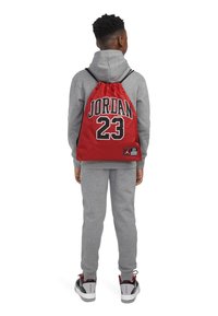 Piros húzózsinóros hátizsák, amelyen feketével van nyomtatva a "JORDAN 23" felirat, szürke kapucnis pulóver és szürke tréningnadr�ág kíséretében, fekete és piros sportcipőkkel párosítva.