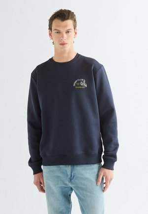 Jeune homme portant un sweat-shirt bleu marine avec le logo « DENIM CLUB WRANGLER » sur la poitrine, associé à un jean bleu clair, debout sur fond blanc.