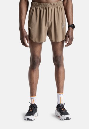 Man draagt beige hardloopshorts, witte sportsokken met oranje strepen, zwart-witte hardloopschoenen en een zwarte smartwatch, staand.