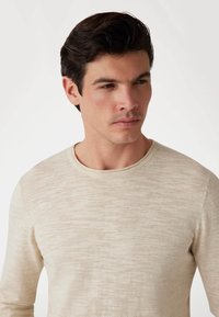 Beige Langarmshirt aus leichtem Stoff mit strukturiertem Muster und Rundhalsausschnitt. Präsentiert an einem Modell mit kurzen dunklen Haaren.