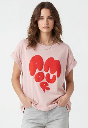 Jeune femme aux cheveux bruns ondulés porte un T-shirt rose clair avec de grandes lettres rouges en forme de cœur formant le mot « AMOUR », mains dans les poches.