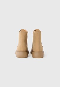 Un par de botas de ante color miel hasta el tobillo con suelas gruesas y cremalleras traseras, vistas desde atrás sobre un fondo blanco liso.