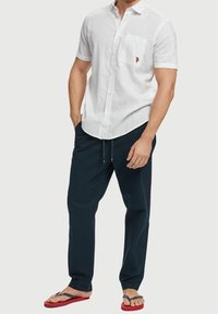 Bílá lněná košile s krátkým rukávem, náprsným kapsičkou a vyšitým logem, doplněná o tmavě modré kalhoty s elastickým páskem a černé žabky s červenými podrážkami.