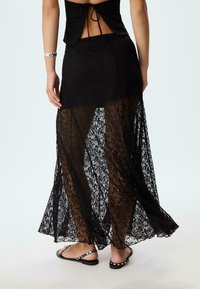Černá krajková maxi sukně s průhledným designem, která má volný siluetu. Tkanina má květinový vzor a je kombinována s plochými sandály.
