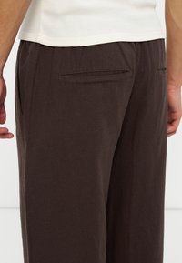 Άτομο που φοράει σκουρόχρωμο καφέ παντελόνι με τσέπες τύπου welt στο πίσω μέρος και λευκό υφασμάτινο πουκάμισο, φωτογραφημένο από τη μέση έως το μέσο του μηρού.