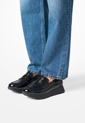 Schwarze Lackleder-Lederstrumpfschuhe mit strukturiertem Finish, niedrigem Keilsohle und dekorativem Riemen auf der Oberseite, kombiniert mit hellblauen Jeans.