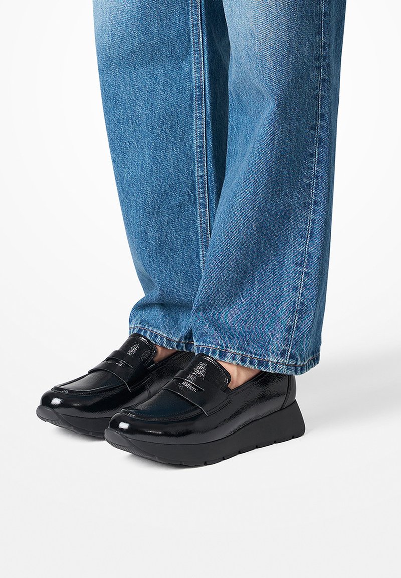 Black patent loafers with a textured finish, low wedge sole, and decorative strap on the upper, paired with light blue denim jeans.