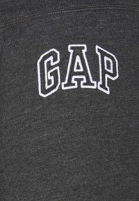 Szara tkanina z białym haftowanym logo "GAP". Materiał wydaje się miękki z delikatną teksturą i klasycznym, casualowym designem.