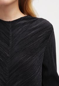 Top negro texturizado con finos pliegues verticales. Presenta un cuello alto y mangas largas, creando una silueta estilizada.