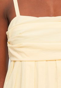 Abito senza maniche in tessuto giallo chiaro con corpetto arricciato e gonna plissettata. Texture liscia, aderente in vita, con spalline semplici.
