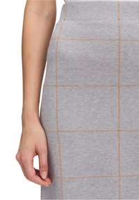 Jupe en tricot gris avec un motif en grille de couleur marron clair, dotée d'une silhouette ajustée et d'une taille haute. Main partiellement visible.