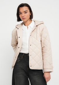 Πουπουλένιο μπεζ σακάκι με κουκούλα, κουμπιά με τρουκς και πλαϊνές τσέπες. Φορεμένο πάνω από λευκό πουκάμισο με γιακά, συνδυασμένο με σκούρο τζιν.