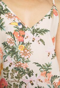 Top senza maniche a fiori con scollatura a V. Tessuto leggero con punti testurizzati, prevalentemente color crema, adornato con fiori vibranti rosa e arancioni e un tucano.