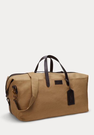 Brown canvas duffel bag with dark leather handles, detachable shoulder strap, zip closure, and leather luggage tag on plain background.