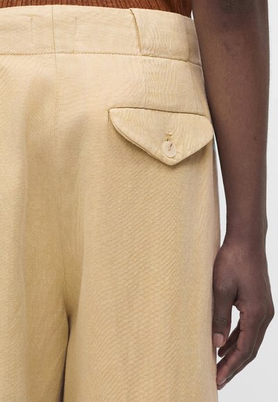 Μπεζ παντελόνι με ψηλή μέση και κουμπωτή τσέπη-καπάκι στο πλάι, φορεμένο με καφέ ριμπ τοπ, εμφανίζοντας το δεξί χέρι στο πλάι.