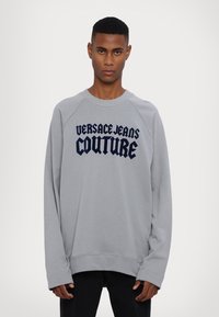 Šedá mikina s námorníckym textom "VERSACE JEANS COUTURE." Navrhnutá s okrúhlym výstrihom a dlhými raglánovými rukávmi pre uvoľnený strih.