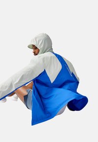 Blau-graue Windjacke mit Kapuze, die ein geometrisches Design und ein leichtes Material aufweist. Die Armbewegung offenbart eine fließende Silhouette.