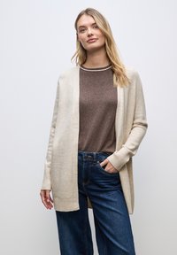 Cardigan beige chiaro con polsini a coste, indossato sopra una maglia marrone. Jeans blu scuro, con vestibilità rilassata. Design semplice e pulito.