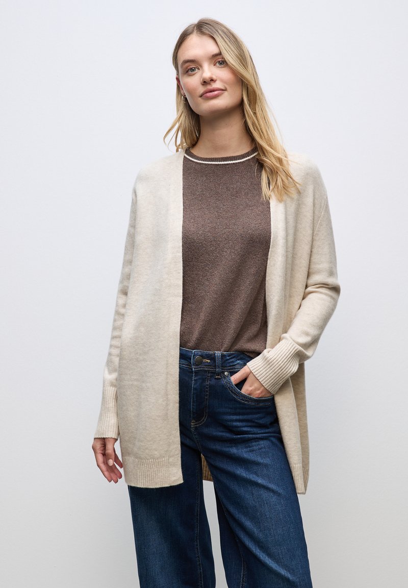 Cardigan beige chiaro con polsini a coste, indossato sopra una maglia marrone. Jeans blu scuro, con vestibilità rilassata. Design semplice e pulito.
