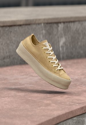 Beige lavtop sneaker med hvide snørebånd og tyk tekstureret sål, vist svævende over en stenet overflade med en grå stenvæg som baggrund.