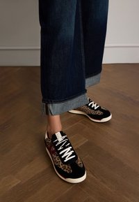 Schwarze Sneaker mit Leopardenmuster und weißen Schnürsenkeln, kombiniert mit dunkelblauen Jeans mit aufgeschlagenem Saum und einem helleren Denim-Akzent am Ende.