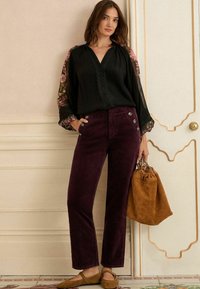 Čierna blúzka s kvetinovými rukávmi, fialové kordové nohavice s tombolovými detailmi a hnedá semišová kabelka. Hnedé baleríny dotvárajú outfit.