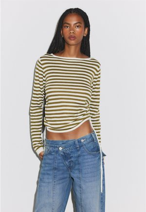 Femme aux longs cheveux tressés portant un haut à manches longues rayé olive et blanc et un jean bleu ample sur un fond uni.