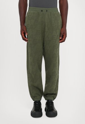 Homme portant un pantalon de jogging texturé vert olive avec des poignets élastiques et des baskets noires à lacets, se tenant contre un fond uni.