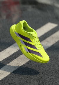 Leuchtend gelber Laufschuh mit violetten Streifen. Leichtes Mesh-Obermaterial, strukturierte Oberfläche und Schnürsenkel. Erhöhte Sohle mit stromlinienförmiger Form.