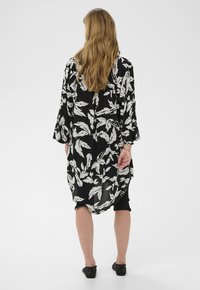 Femme aux longs cheveux blonds portant une tunique ample noire et blanche à motif de feuilles, un pantalon noir raccourci et des chaussures noires, debout de dos.