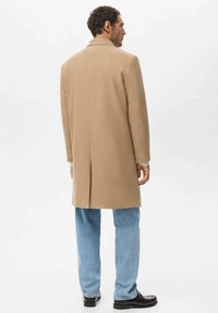 Abrigo de lana beige con corte recto, abertura trasera y solapa con muesca, combinado con vaqueros de mezclilla azul claro y zapatos negros.