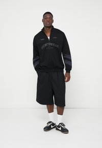 Felpa nera a collo alto con zip e testo "SPORTSWEAR", abbinata a pantaloni corti neri. Presenta accenti grigi a righe sulle maniche e calze bianche.