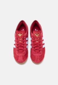 Κόκκινα σουέντ αθλητικά παπούτσια με υφή, που διαθέτουν λευκές ρίγες, χρυσά διακοσμητικά λογότυπα και σόλα από καουτσούκ καραμέλας.