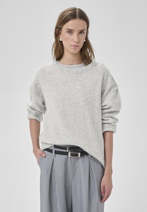 Kvinde med skulderlangt brunt hår, iført perleøreringe og lysegrå sweater, stoppet ned i bredbenede grå bukser med høj talje og dobbelt sort bælte.