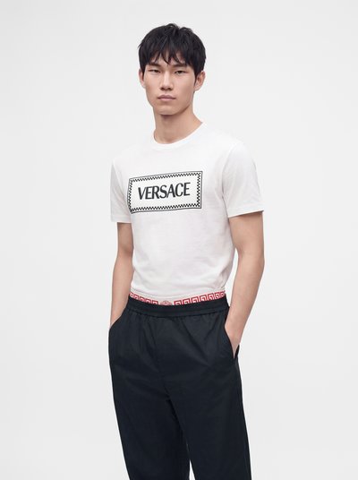Νέος άνδρας που φοράει λευκό t-shirt Versace και μαύρο παντελόνι με τα χέρια στις τσέπες, στέκεται μπροστά σε φωτεινό απλό φόντο.