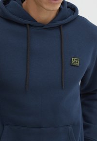 Marinblå hoodie av mjukt tyg, med en framficka, dragsko med svarta spetsar och en liten grön logopatch på bröstet.