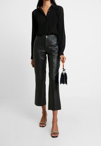 Camicia nera a maniche lunghe plissettata, abbinata a pantaloni in pelle nera a vita alta, accentuata da una borsa piccola e tacchi aperti con cinturini.
