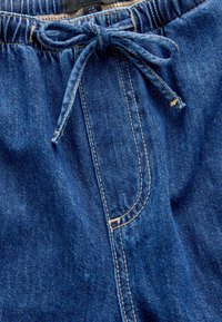 Modré rifľové nohavice s elastickým pásom so sťahovacou šnúrkou, prešívanými detailmi a predným vreckom. Textúra je hladká a mierne drsná.