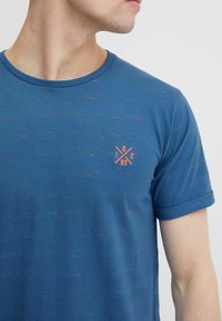 Tricou scurt cu mânecă albastră dintr-un material texturat cu mici nuanțe de culoare. Are un logo portocaliu mic pe piept. Decolteu rotund.