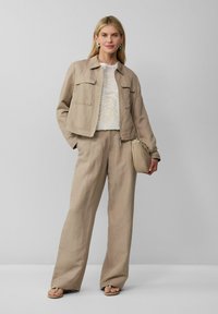 Femme portant une veste beige et un pantalon large, un haut à motifs blancs, tenant un sac à main beige, debout sur un fond gris uni.