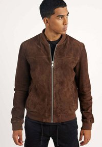 Veste bomber en daim marron avec fermeture éclair, poignets côtelés et texture lisse. Présente un design simple et sans motifs visibles.