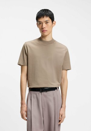 Junger Mann mit kurzen schwarzen Haaren, der ein beiges kurzärmliges Hemd und hellgraue hoch taillierte Hosen mit einem schwarzen Gürtel trägt.