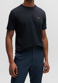 Černé bavlněné tričko s kulatým výstřihem, na hrudi s malým zlatým logem "BOSS". V kombinaci s námořnickými kalhotami.